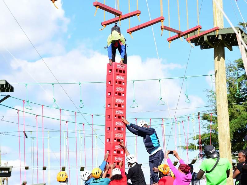 A climber on a safety rope stands on a tall stack of red crates while teammates pass crates up